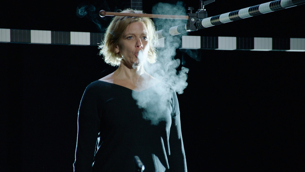 Eine Frau steht vor schwarzem Hintergrund und atmet aus. Ihr Atmen ist im Gegenlicht als Dampfwolke zu erkennen.