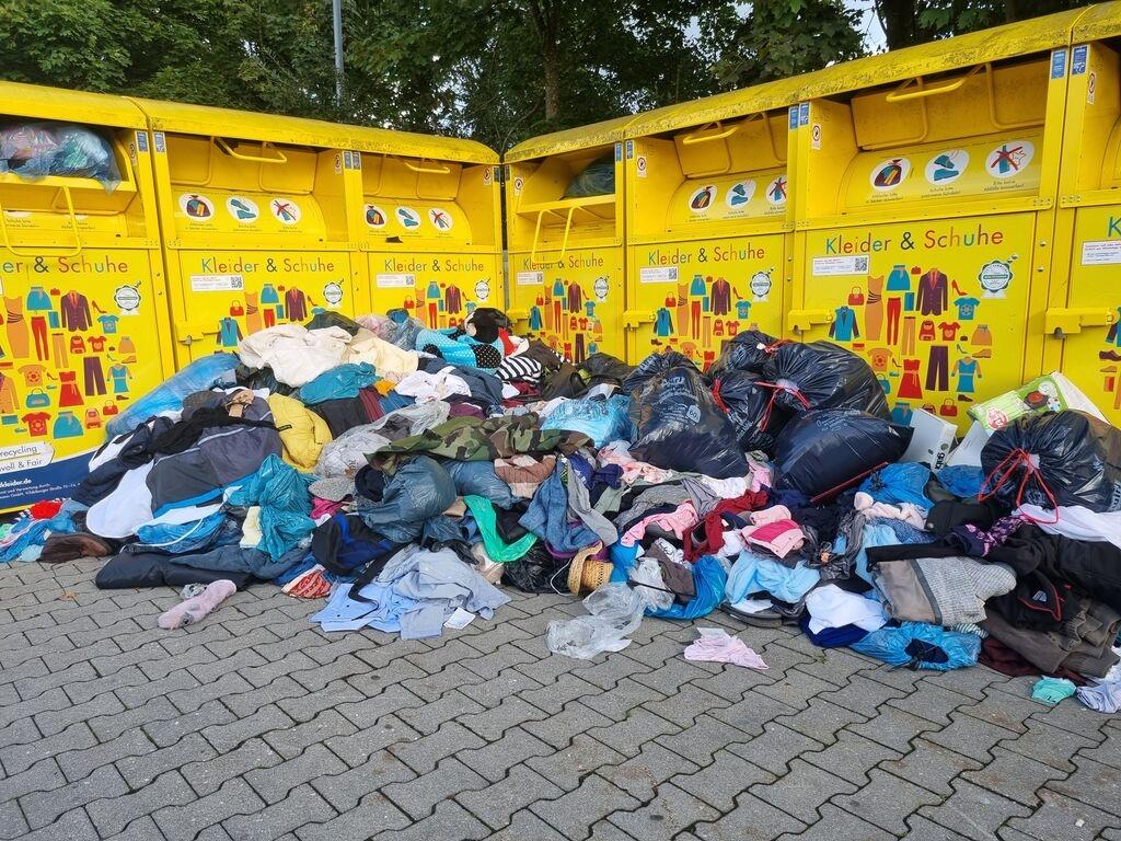 (Symbolbild) Altkleider liegen in Haufen vor Containern auf dem Boden.