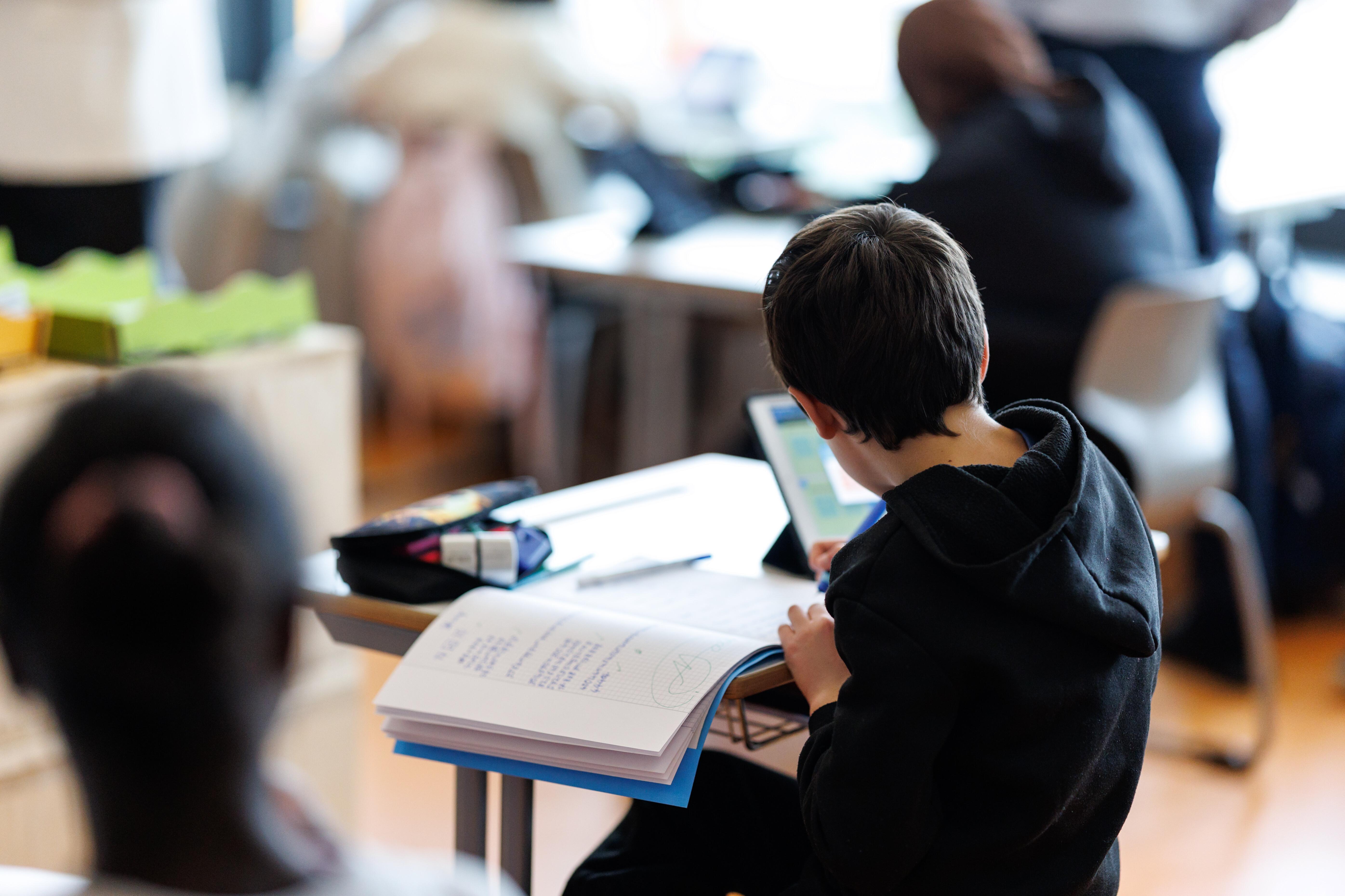(Symbolbild) Ein Schüler sitzt mit Heft und Tablet an einem Tisch in der Schule