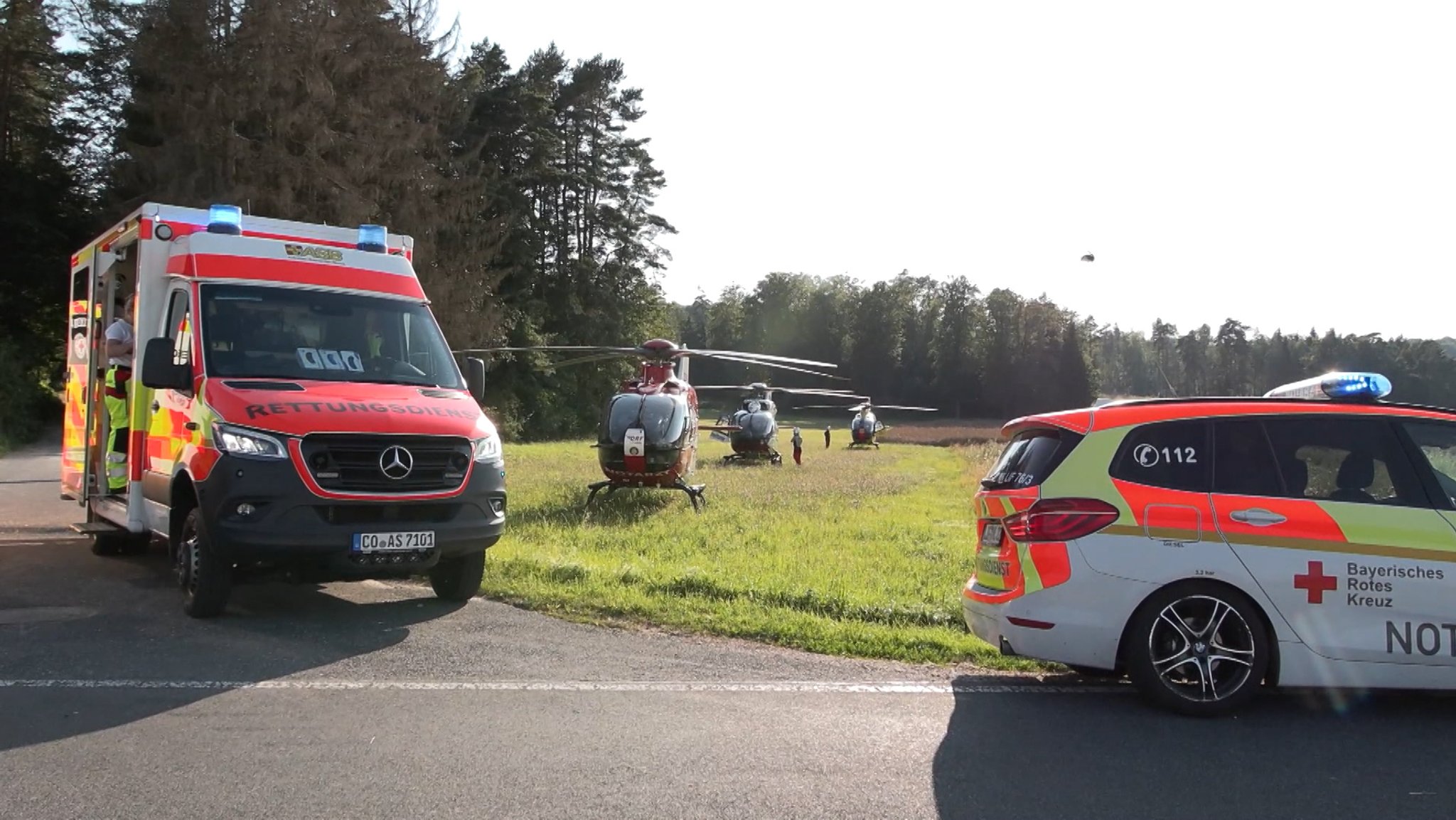 Auf einer Landstraße stehen zwei Rettungswagen, auf der Wiese daneben drei Hubschrauber, dazwischen bewegen sich Rettungskräfte. Nach einer Fahrt in einem überhitzten Bus sind fünf Jugendliche mit Verdacht auf schweren Hitzschlag ins Krankenhaus gebracht worden. 