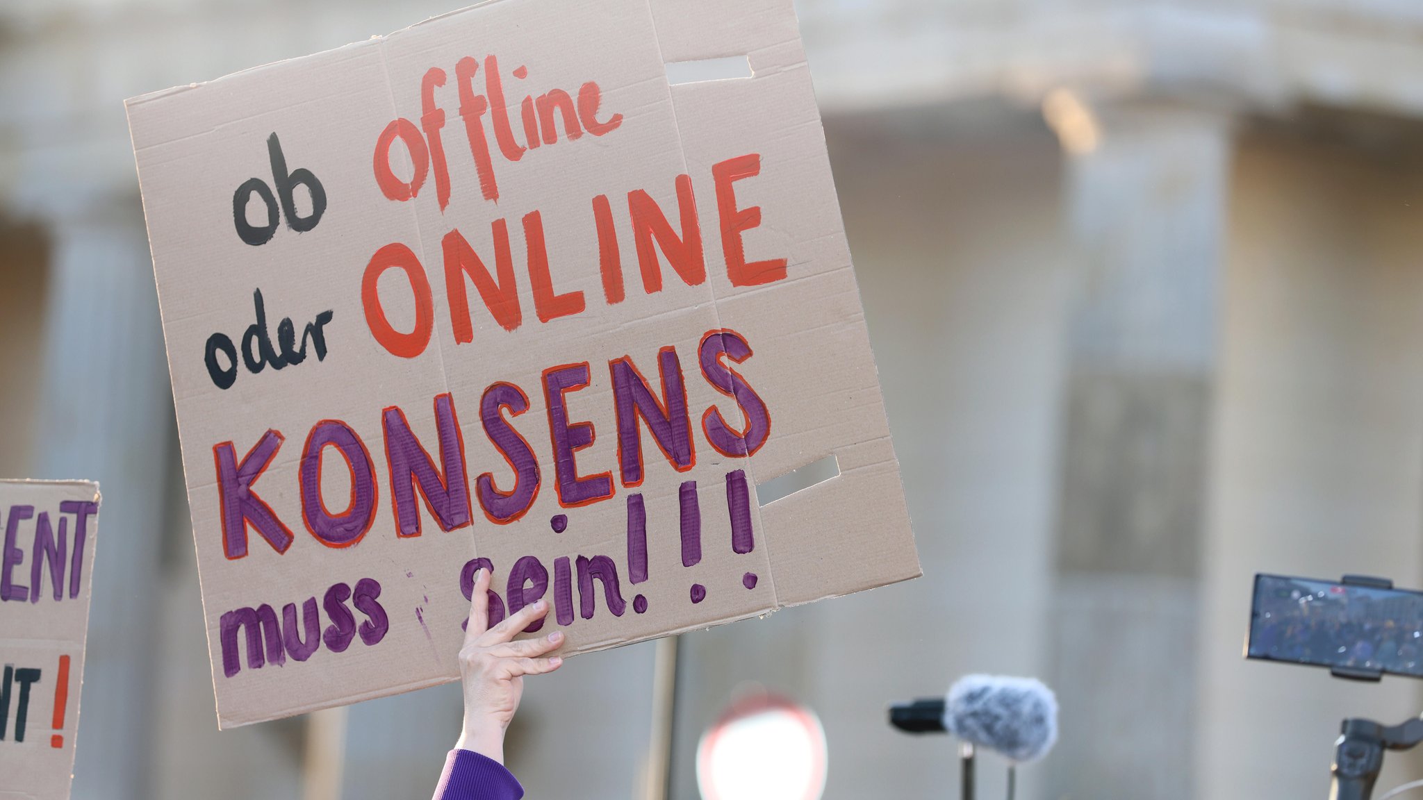 (Symbolbild) Ein Demoplakat aus Pappe wird in die Luft gehalten, auf dem steht: "Ob offline oder Online - Konsens muss sein!!!"