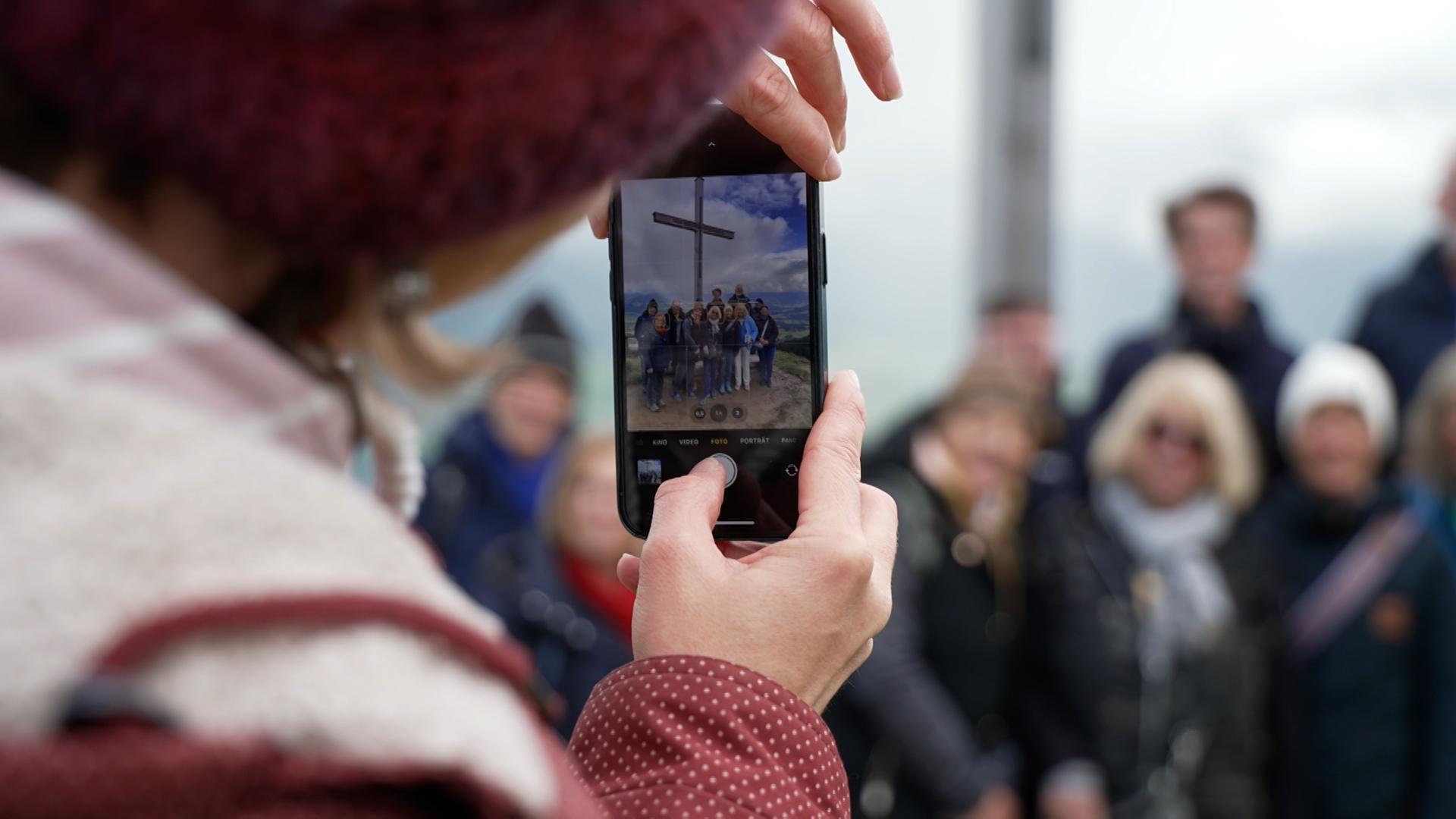 Eine Gruppe steht vor dem Gipfelkreuz am Ofterschwanger Horn. Im Vordergrund fotografiert eine Person mit dem Handy.