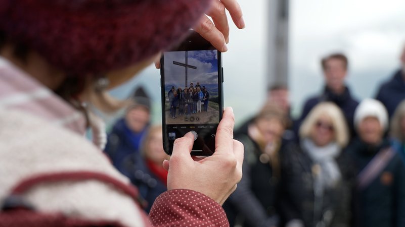 Eine Gruppe steht vor dem Gipfelkreuz am Ofterschwanger Horn. Im Vordergrund fotografiert eine Person mit dem Handy. | Bild: BR/German Gross Eine Gruppe steht vor dem Gipfelkreuz am Ofterschwanger Horn. Im Vordergrund fotografiert eine Person mit dem Handy.
