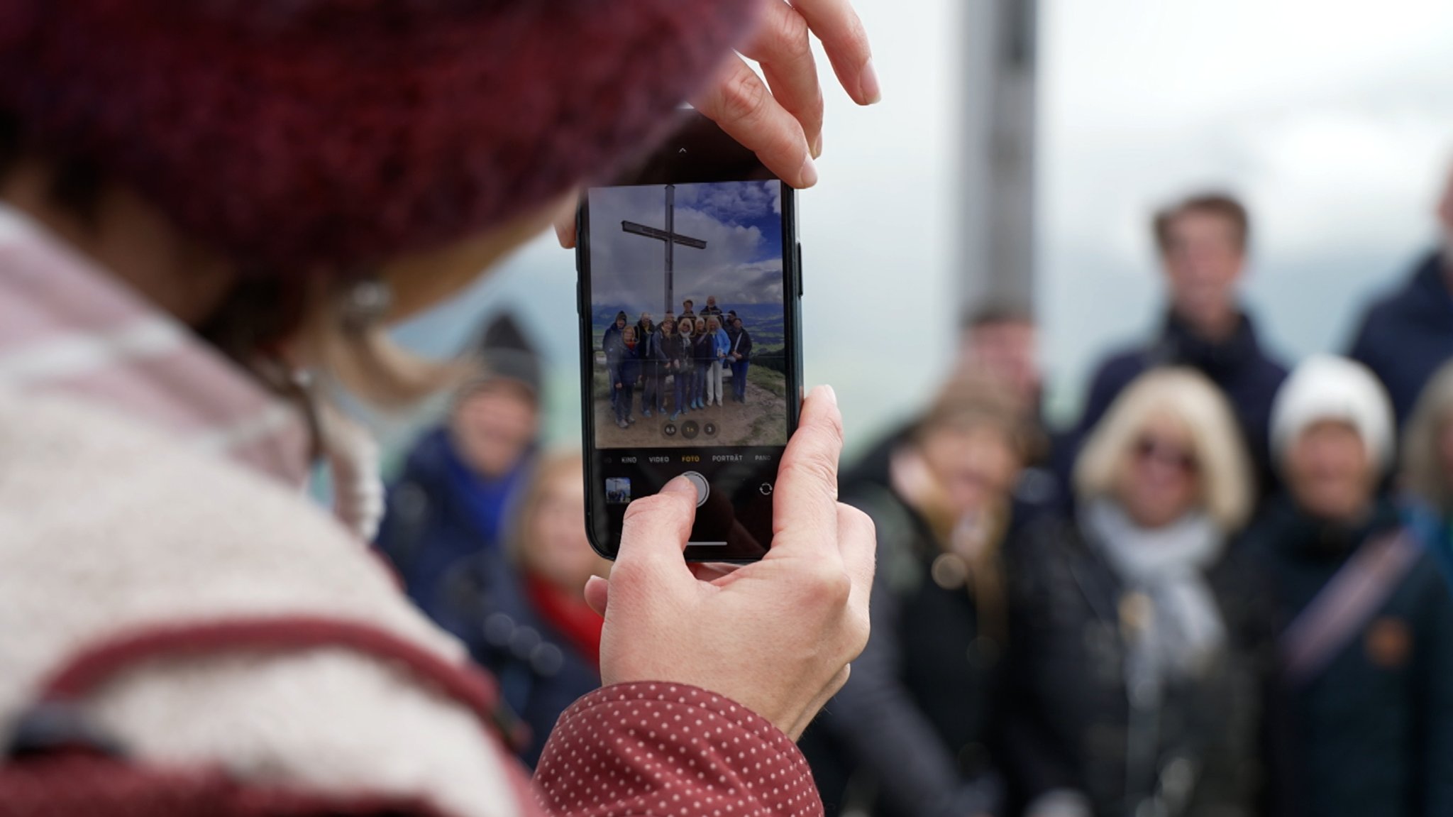 Eine Gruppe steht vor dem Gipfelkreuz am Ofterschwanger Horn. Im Vordergrund fotografiert eine Person mit dem Handy.