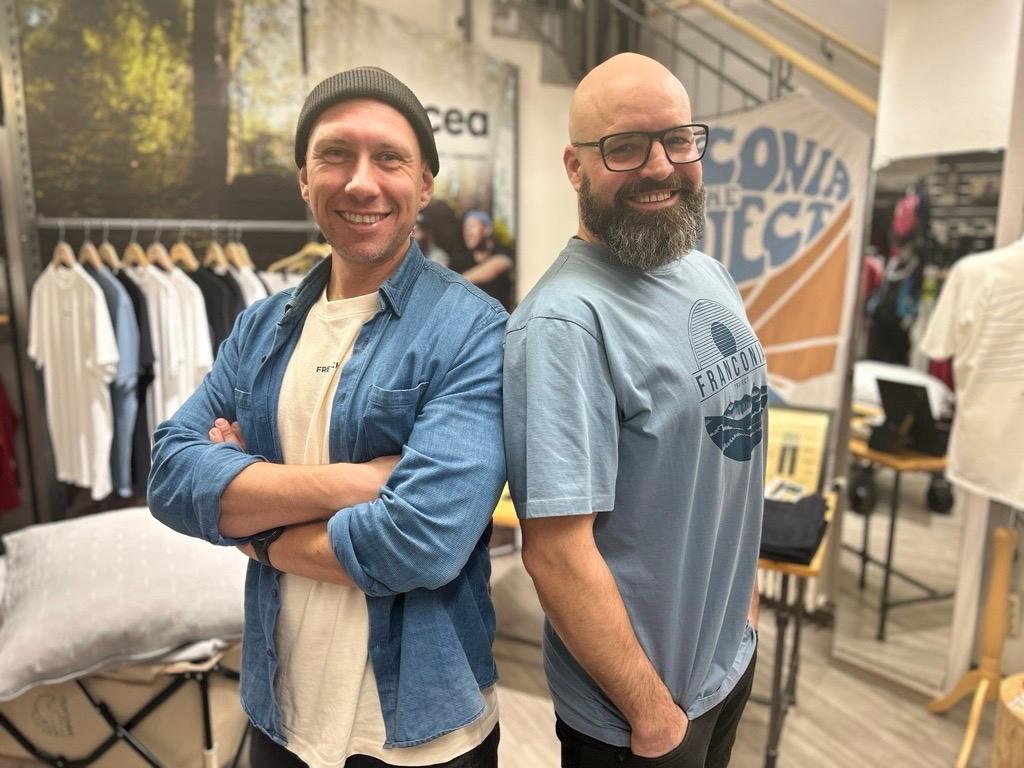 Zwei Männer stehen Rücken an Rücken, dahinter ein Kleiderständer mit Hemden. 