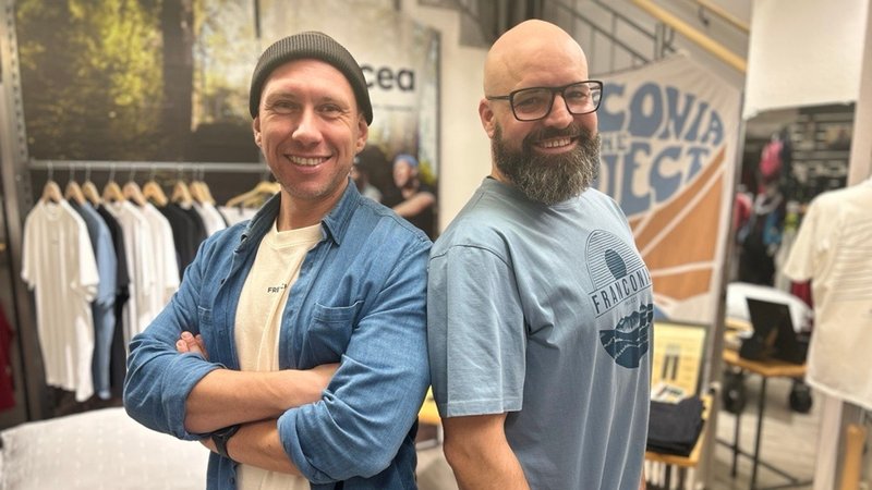 Zwei Männer stehen Rücken an Rücken, dahinter ein Kleiderständer mit Hemden. | Bild: BR/Kristina Kreutzer Zwei Männer stehen Rücken an Rücken, dahinter ein Kleiderständer mit Hemden.