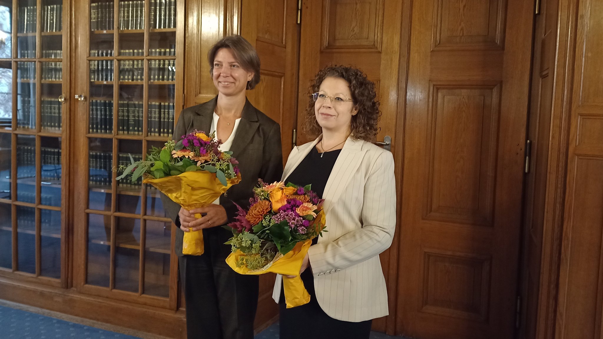 Zwei Frauen stehen mit Blumensträußen in einer Bibliothek.