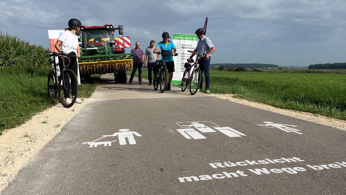 "Rücksicht macht Wege breit"-Schriftzug auf einem Radweg. Im Hintergrund Radfahrer und Landwirte mit einem Traktor.