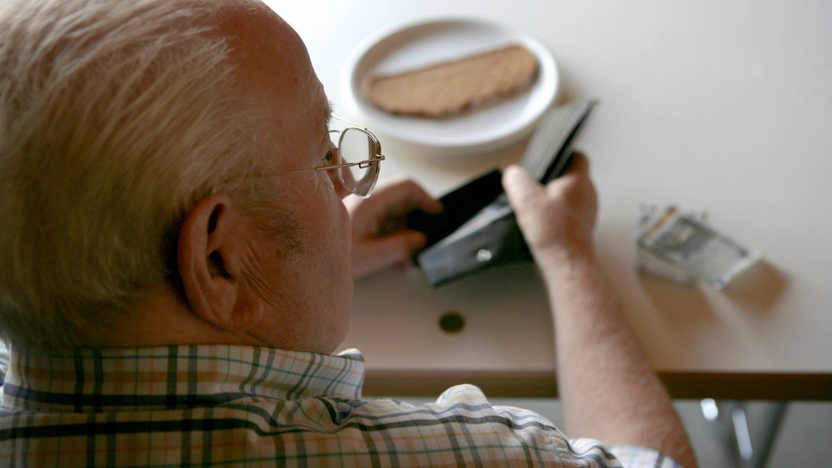 Rentner sieht in eine leere Geldbörse, dahinter eine trockene Scheibe Brot (gestelltes Symbolbild)