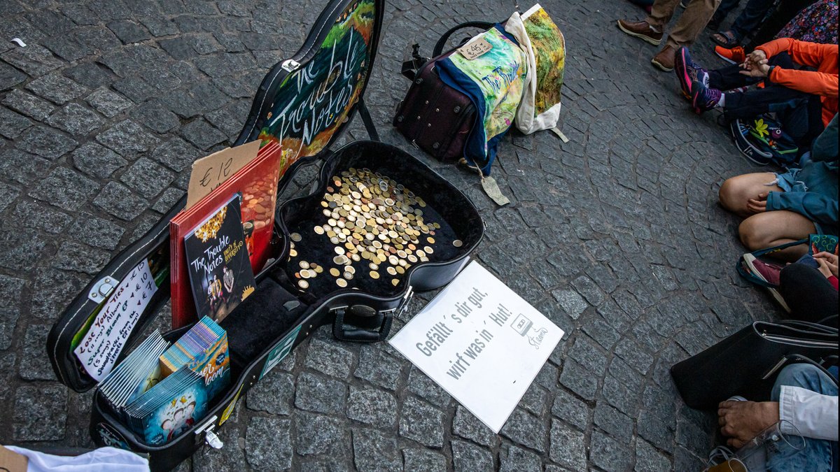 Ein Gitarrenkoffer, darin Münzen, davor Schilder der Band und Merch.