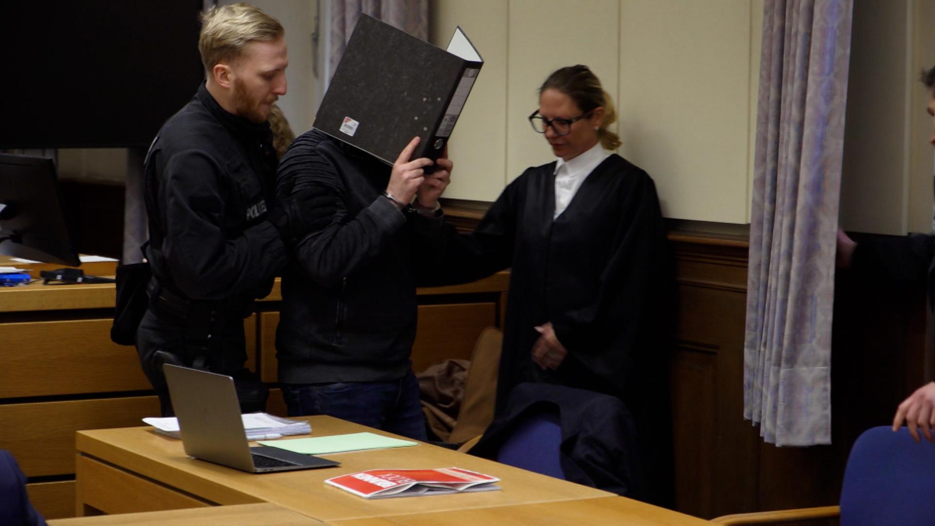 Der heute 22-jährige Angeklagte betritt den Gerichtsssaal und verhüllt dabei sein Gesicht mit einem Ordner.