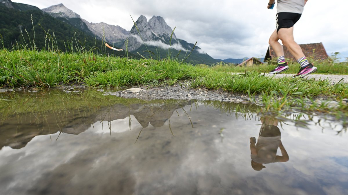 Ein Jogger spiegelt sich zusammen mit den Gipfeln des Wettersteingebirges in einer Regenpfütze.