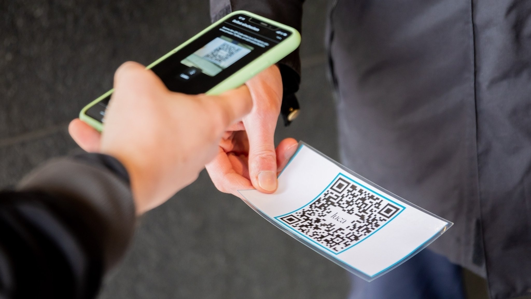 Eine Person scannt am Eingang eines Bekleidungsgeschäftes vor einem Sicherheitsmitarbeiter mit der Luca-App einen QR-Code. Die App dient der Datenbereitstellung für eine mögliche Kontaktpersonennachverfolgung.