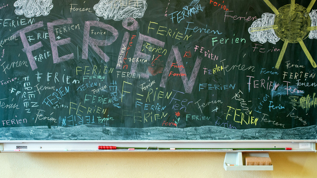 Schultafel in einem Klassenzimmer, auf der ,it bunten Kreiden viele Male das Wort Ferien angeschrieben steht