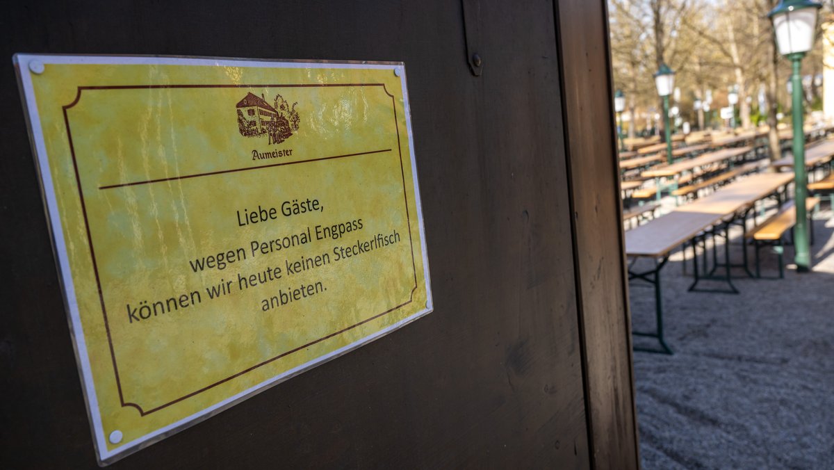 Ein Schild mit der Aufschrift "Liebe Gäste, wegen Personal Engpass können wir heute keinen Steckerlfisch anbieten" hängt in einem Biergarten.