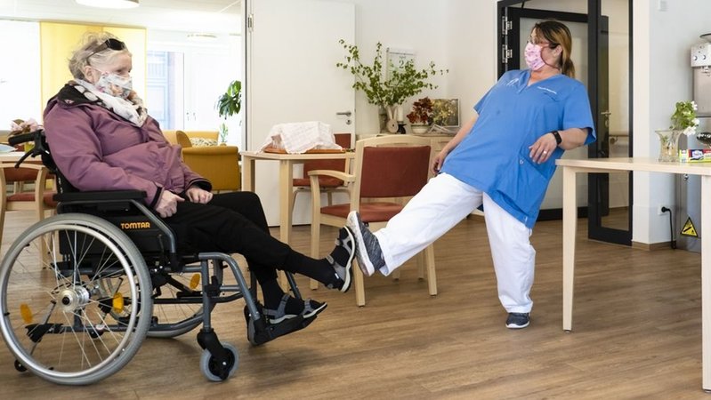 Eine Bewohnerin einer Pflegeeinrichtung begrüßt eine Pflegerin mit dem Fuß.