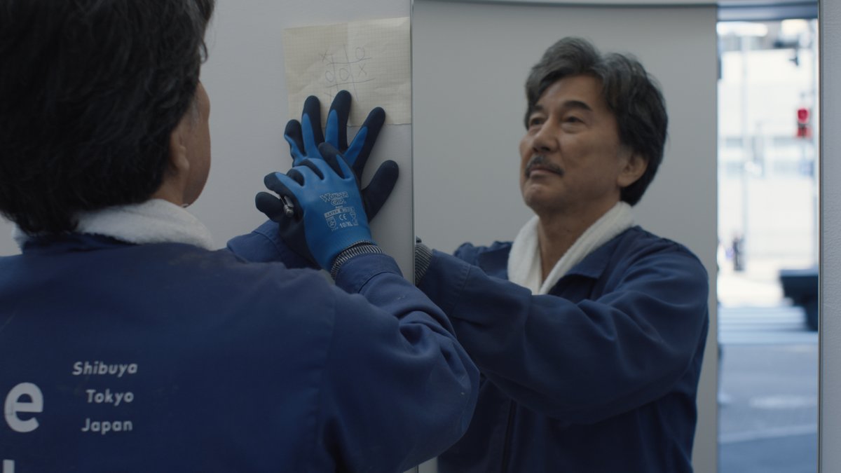 Japanischer Reinigungsarbeiter mit Gummihandschuhen vorm Spiegel in öffentlicher Toilette