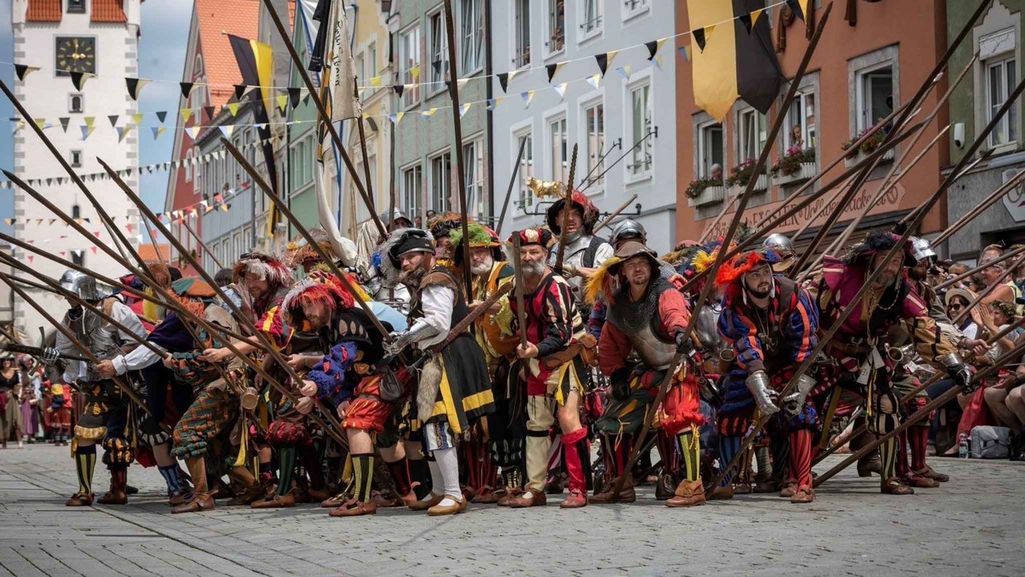 Männer in traditionellen Gewändern aus dem Jahre 1500 halten Speere und Spieße in die Luft.