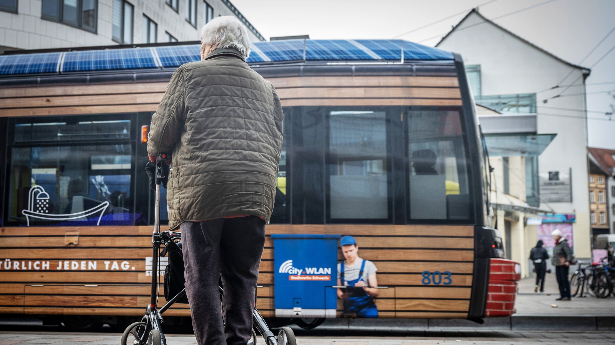 Eine ältere Person mit Rollator steht an einer Straßenbahnhaltestelle und wartet auf die Straßenbahn. 