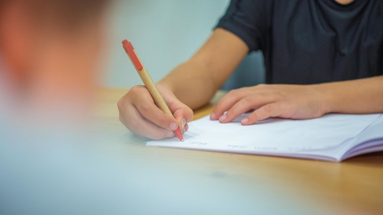 Ein Grundschüler sitzt an einem Tisch und schreibt mit einem Kugelschreiber in sein Heft. | Bild:picture alliance/dpa | Mohssen Assanimoghaddam