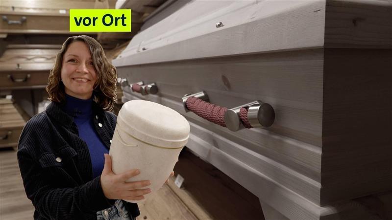 Nadine hält eine Urne in der Hand und steht vor Särgen