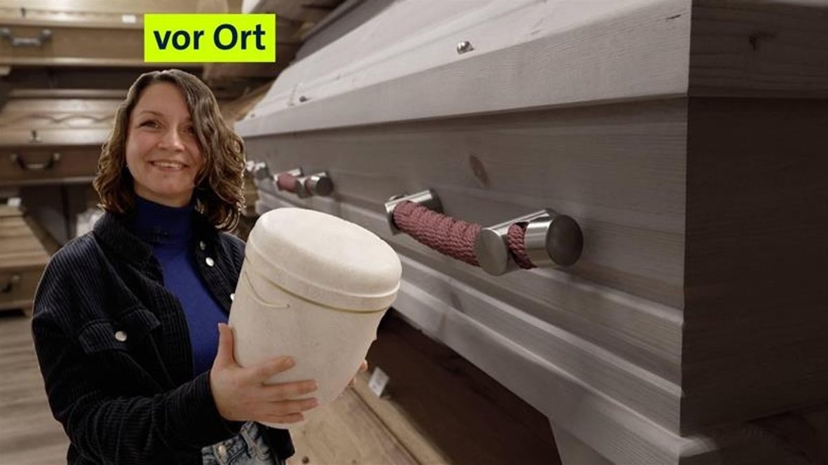 Nadine hält eine Urne in der Hand und steht vor Särgen | Bild: BR24 Nadine hält eine Urne in der Hand und steht vor Särgen