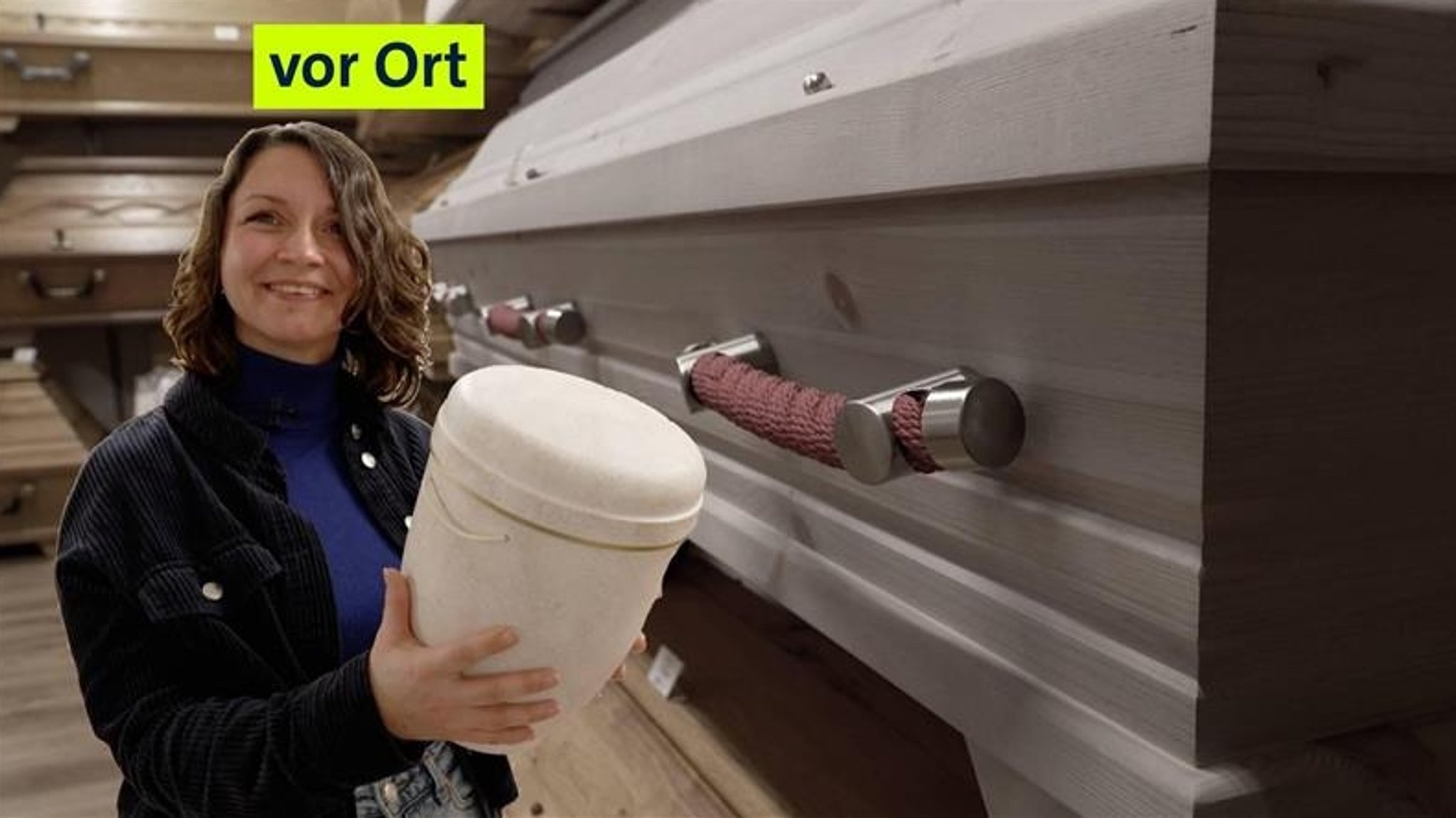 Nadine hält eine Urne in der Hand und steht vor Särgen