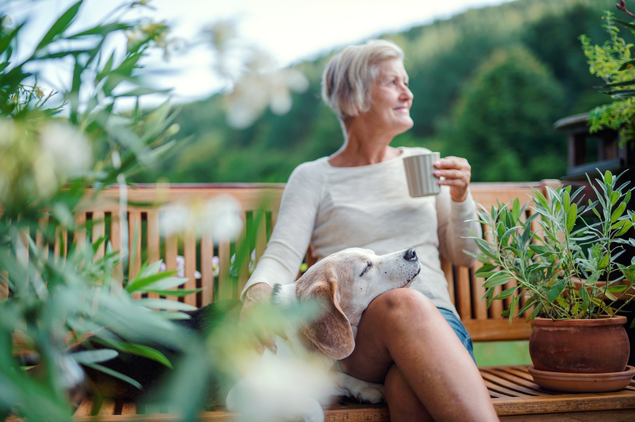 Eine Rentnerin sitzt an einem Sommertag mit ihrem Hund und einer Tasse Kaffee auf der Terrasse.