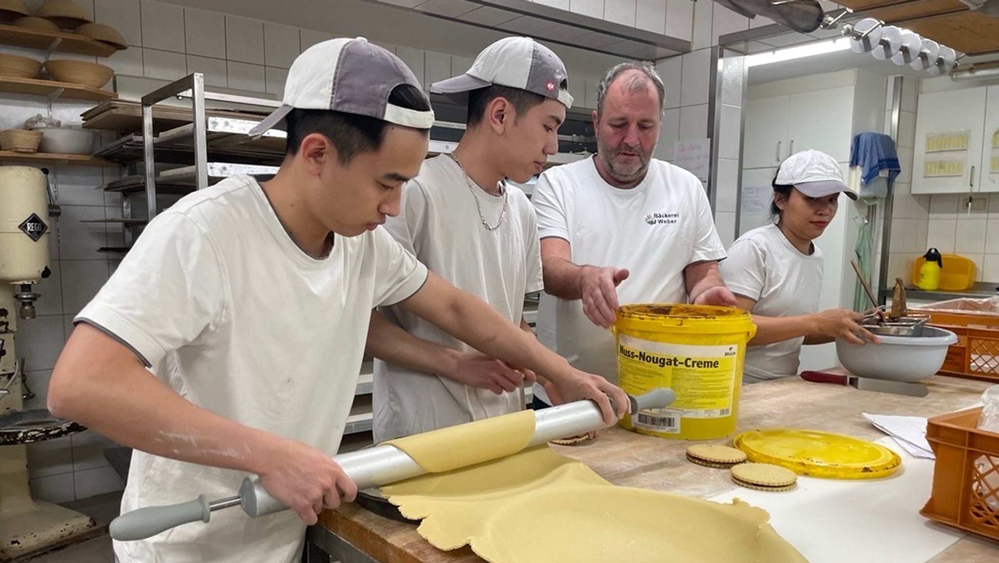 Bäckereichef Erwin Weber leitet seine vietnamesischen Auszubildenden an, einer von ihnen rollt Teig aus.