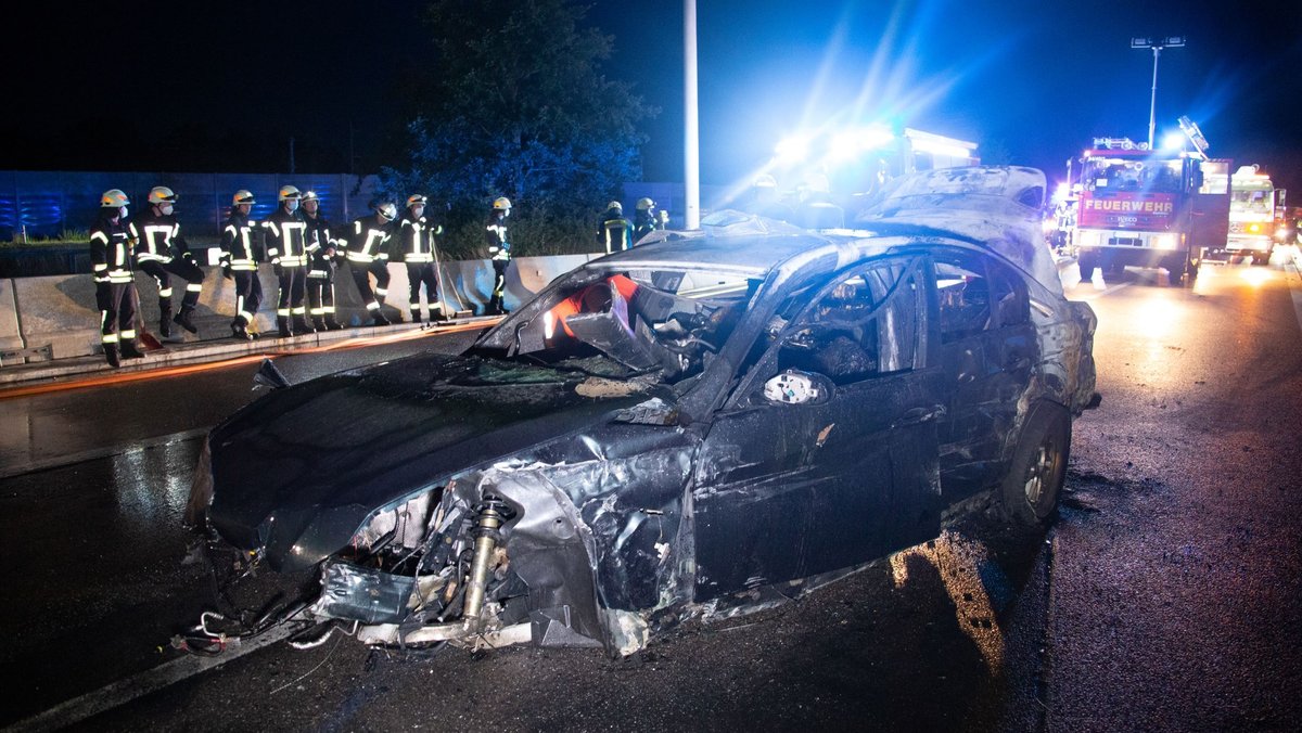 Komplett zerstörtes Auto auf der nächtlichen Autobahn, dahinter Feuerwehrleute.