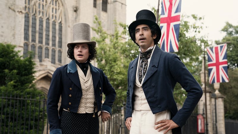 Zwei Männer in historischen Kostümen stehen vor einer Kirche mit britischen Flaggen: Szene aus "David Copperfield"