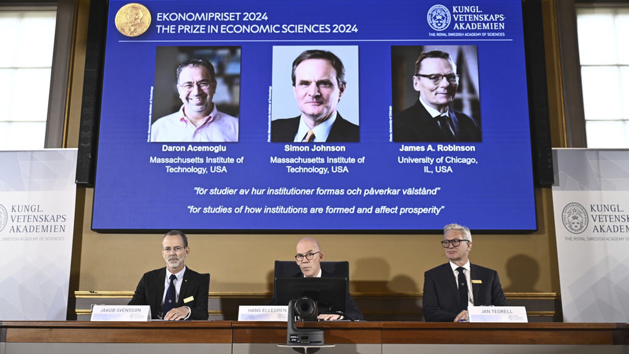 14.10.2024, Schweden, Stockholm: Der ständige Sekretär der Akademie der Wissenschaften, Hans Ellegren (M), Jakob Svensson (l) und Jan Teorell von der Nobelversammlung verkünden die Gewinner des Nobelpreises für Wirtschaftswissenschaften - das sind Daron Acemoglu, Simon Johnson und James A. Robinson (auf dem Bildschirm), während eines Pressetermins in der Königlich Schwedischen Akademie der Wissenschaften in Stockholm. Foto: Christine Olsson/TT News Agency/AP +++ dpa-Bildfunk +++