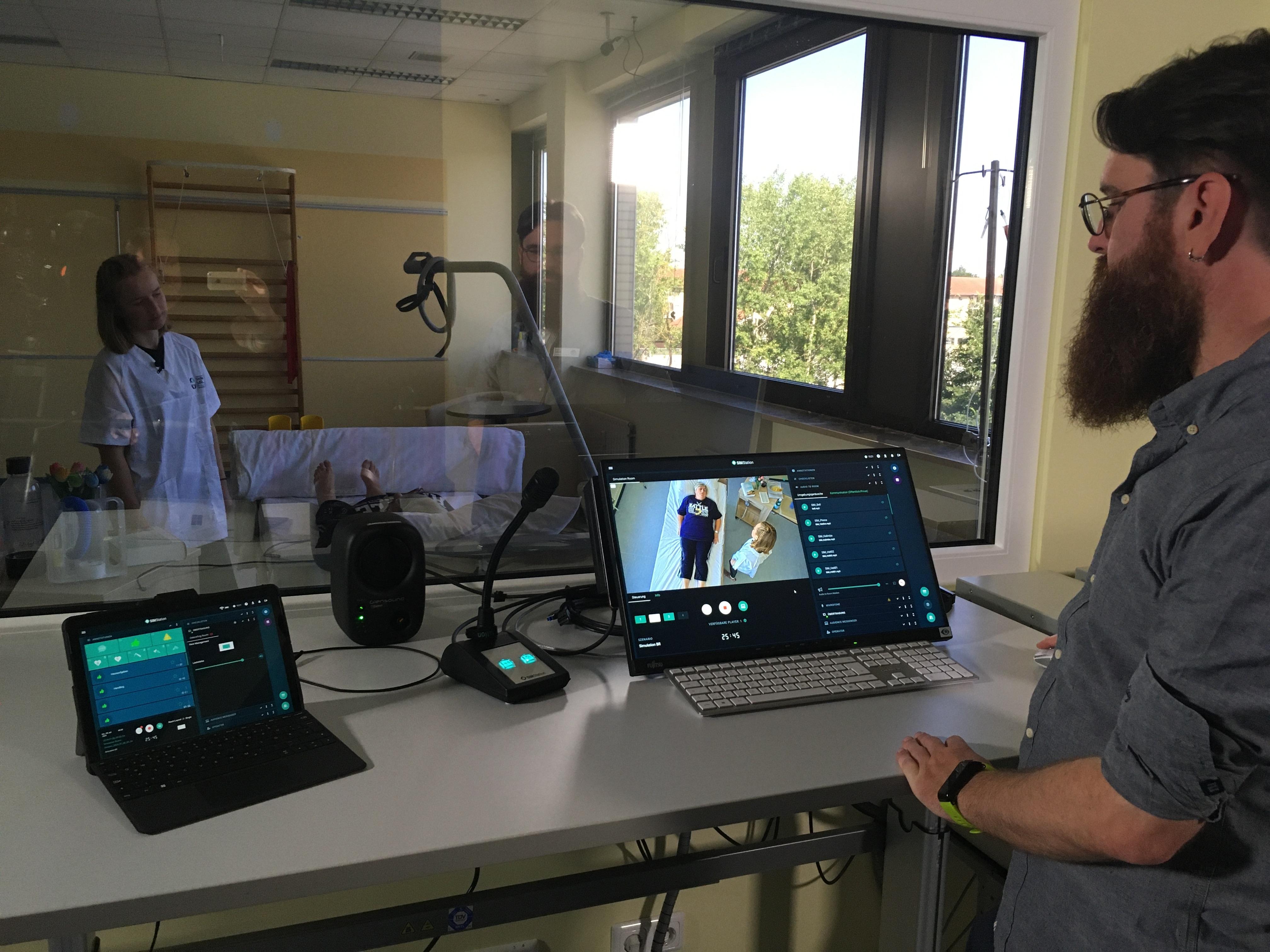 Ein Mann mit Bart und Brille steht an einem Bildschirm und sieht durch ein Fenster in ein Krankenzimmer.