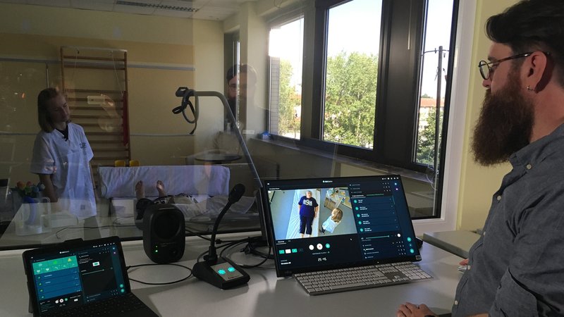 Ein Mann mit Bart und Brille steht an einem Bildschirm und sieht durch ein Fenster in ein Krankenzimmer. | Bild: BR24 / Tobias Burkert Ein Mann mit Bart und Brille steht an einem Bildschirm und sieht durch ein Fenster in ein Krankenzimmer.