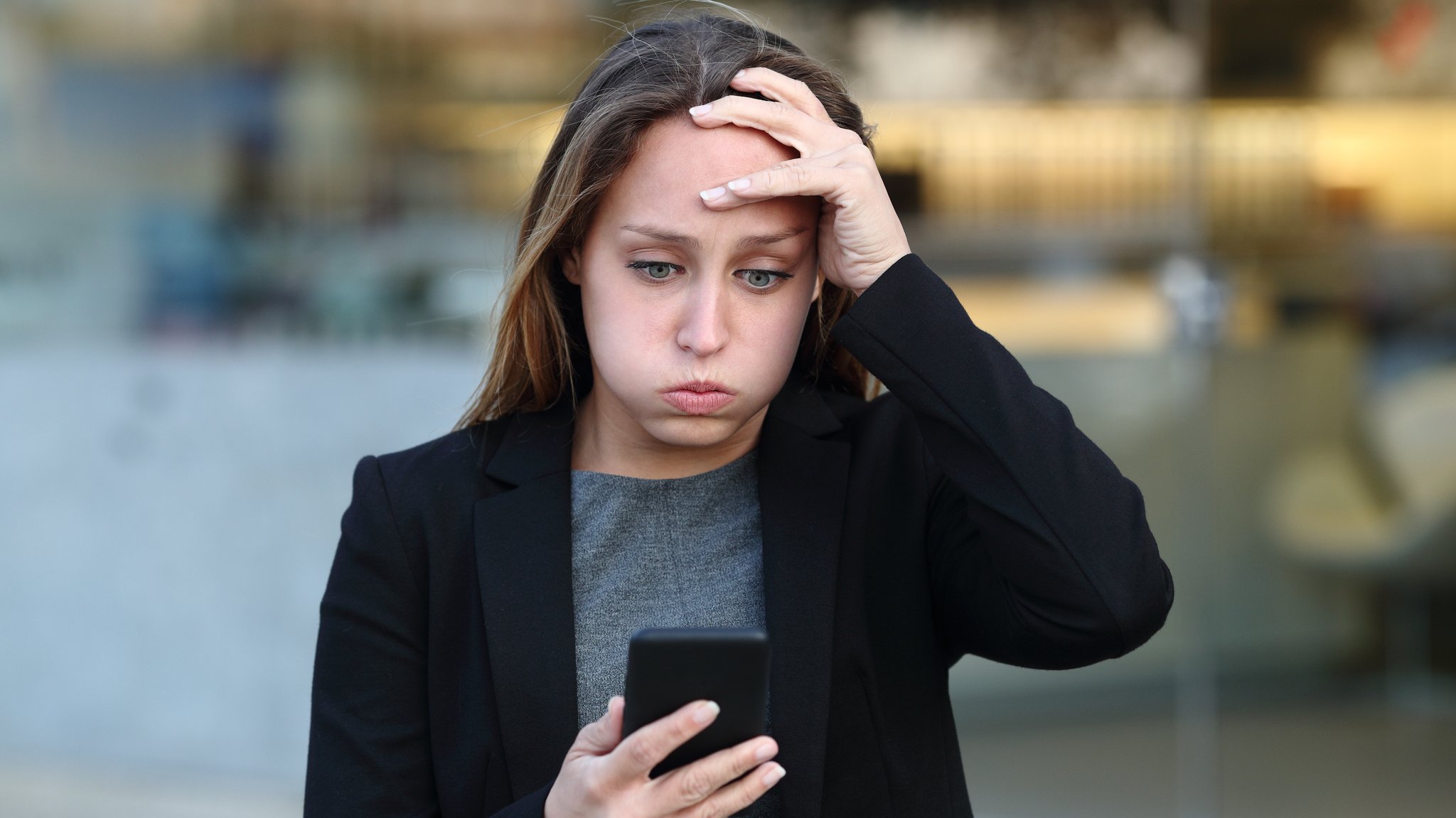 Eine junge Frau schaut auf ihr Handy und bläst die Luft aus und packt sich mit einer Hand an den Kopf.