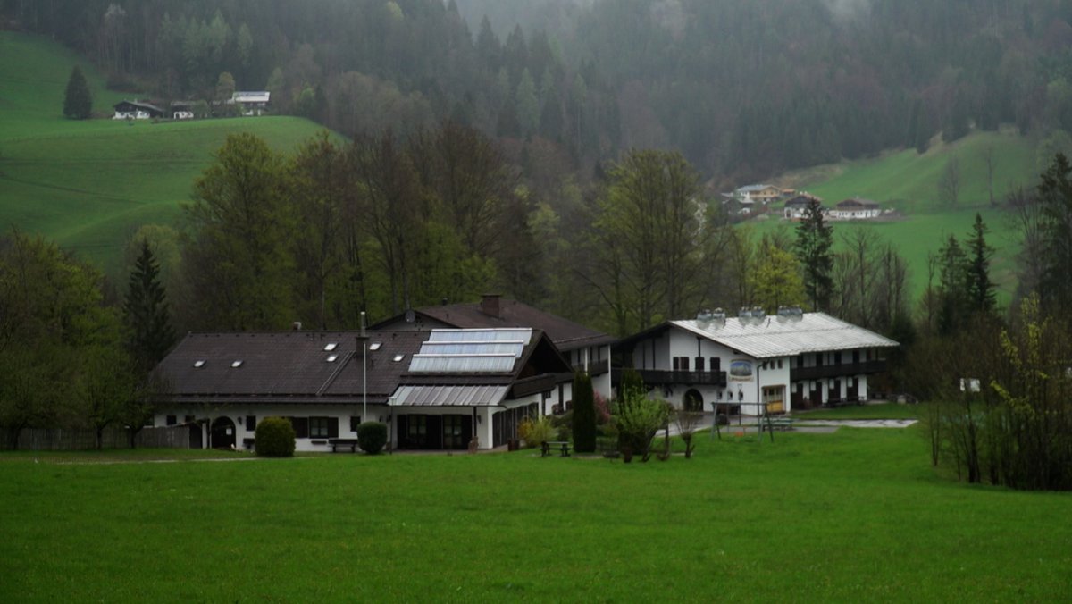 Zwei weiße Häuser im bayerischen Stil liegen von Bäumen eingerahmt auf einer grünen Wiese.