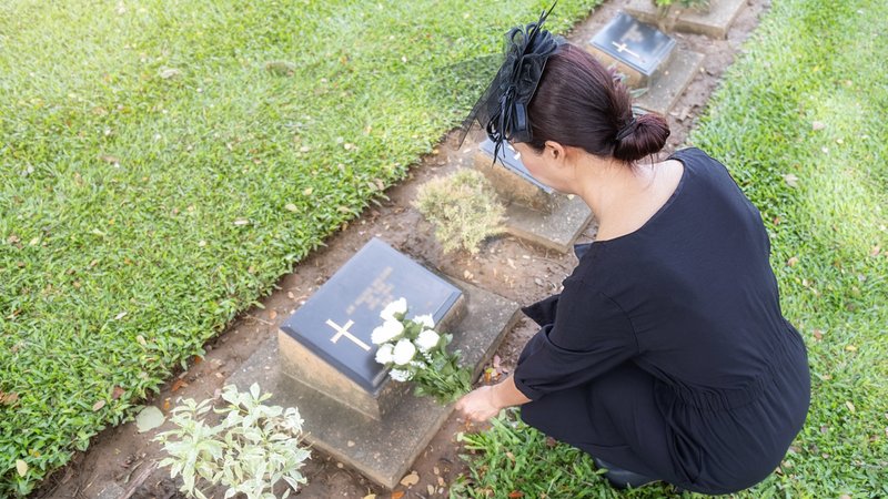 Eine Frau legt Blumen auf einen kleinen Grabstein. | Bild: stock.adobe.com/PIC SNIPE Eine Frau legt Blumen auf einen kleinen Grabstein.
