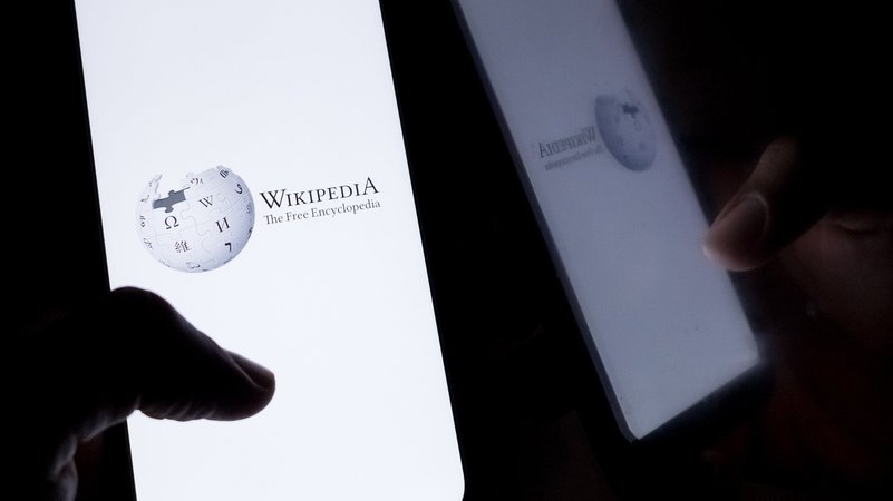 Eine Hand hält ein Handy vor schwarzem Hintergrund. Auf dem Bildschrim ist das Wikipedia-Logo zu sehen. | Bild: picture alliance / Nikolas Kokovlis Eine Hand hält ein Handy vor schwarzem Hintergrund. Auf dem Bildschrim ist das Wikipedia-Logo zu sehen.