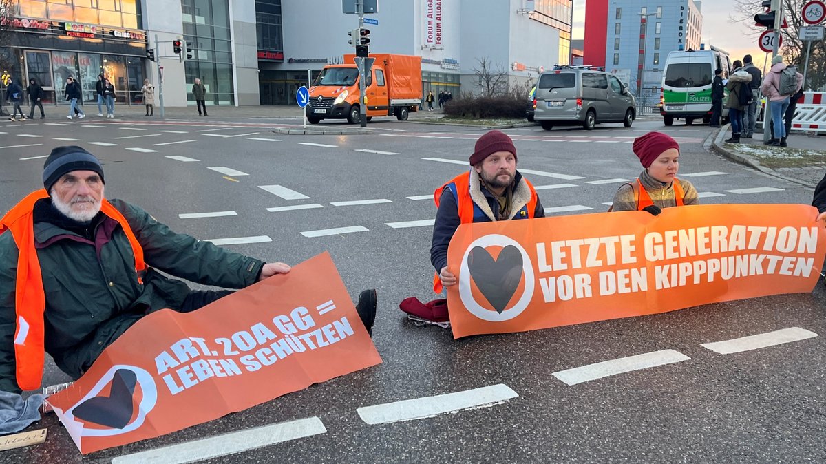 Drei Klimaaktivisten der "Letzten Generation" sitzen  auf einer Straße vor der Forum Allgäu in Kempten und blockieren sie.