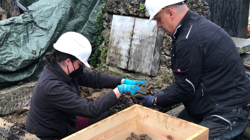 Ein Grab wird von zwei Mitarbeiter ausgehoben. Wo einst Gebeine lagen, soll bald eine Wasserleitung durchführen. | Bild: BR Ein Grab wird von zwei Mitarbeiter ausgehoben. Wo einst Gebeine lagen, soll bald eine Wasserleitung durchführen.