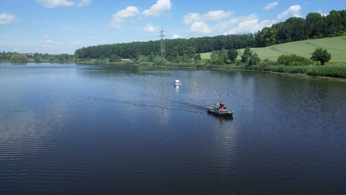 Auf einem großen See, an dessen Ufer grüne Bäume und Felder zu sehen sind, ist ein Boot unterwegs. 