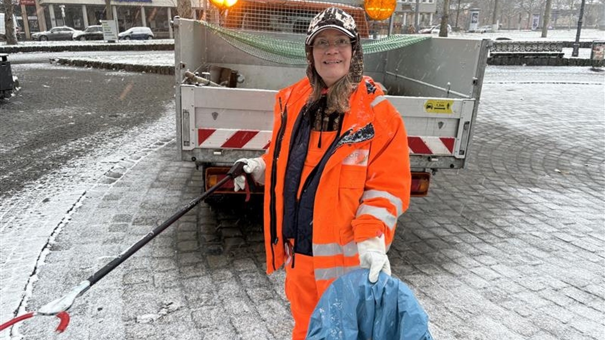 Eine Frau in orangenen Klamotten mit Reflektoren steht mit einem Müllsack und einer Greifzange in der Stadt. Hinter ihr steht ein Fahrzeug der Straßenreinigung. Auf dem Boden liegt etwas Schnee. 