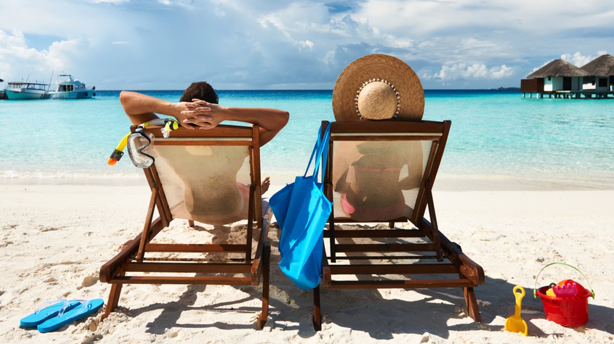 Symbolbild: Ein Pärchen in Liegestühlen am Strand und schaut auf türkisblaues Meer. In Griechenland werden Strandliegen nun beschränkt. 
