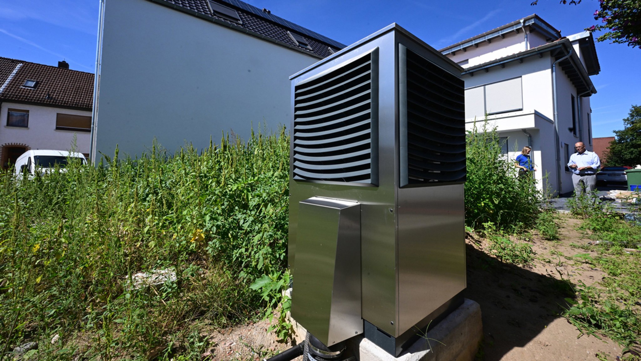 Vor einem neu gebauten Wohngebäude ist im Garten eine Wärmepumpe zum Heizen des Gebäudes in Betrieb. 