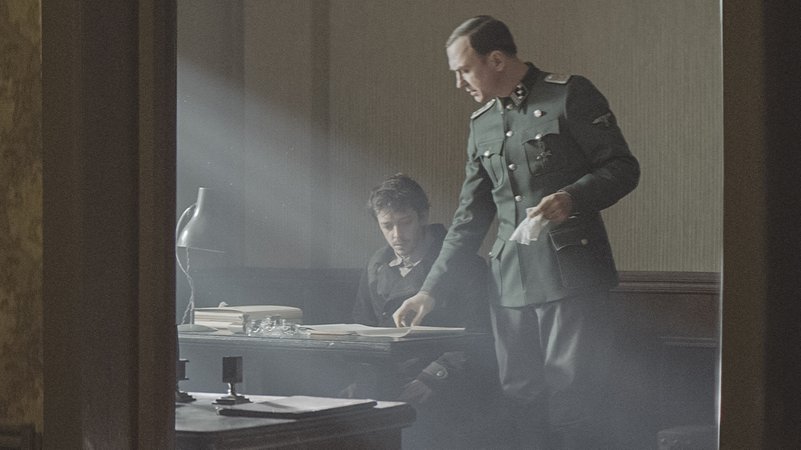 Ein ängstlich guckender Mann sitzt vor einem Tisch. Ein Mann in Nazi-Uniform beugt sich über sein Buch: Szene aus dem Film "Persischstunden"