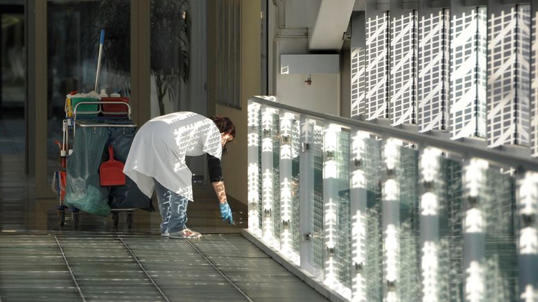 Eine Frau im weißen Kittel mit Putzwagen säubert den Boden eines langen Gangs in einem Betriebsgebäude | Bild:picture alliance / SvenSimon | FrankHoermann/SVEN SIMON