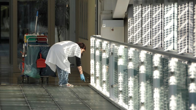 Eine Frau im weißen Kittel mit Putzwagen säubert den Boden eines langen Gangs in einem Betriebsgebäude