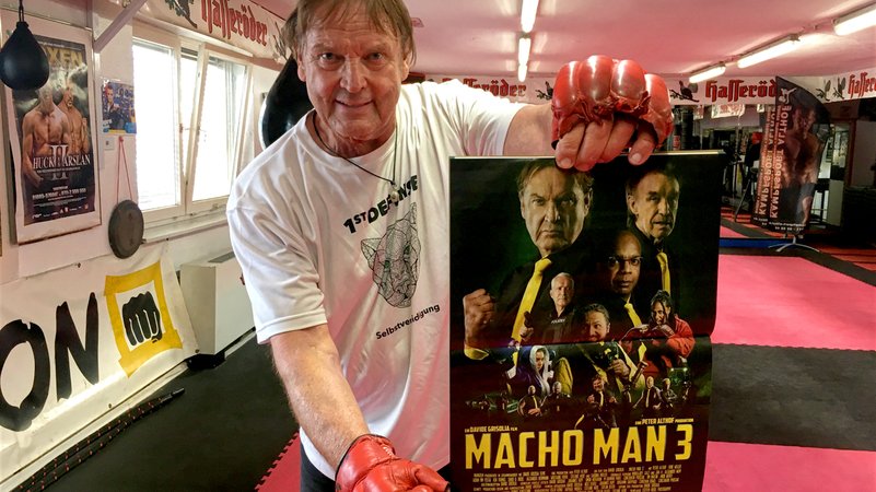 Peter Althof, Personenschützer, hält ein Plakat zu Macho Man 3 in die Kamera. | Bild: BR24 / Tobias Burkert Peter Althof, Personenschützer, hält ein Plakat zu Macho Man 3 in die Kamera.
