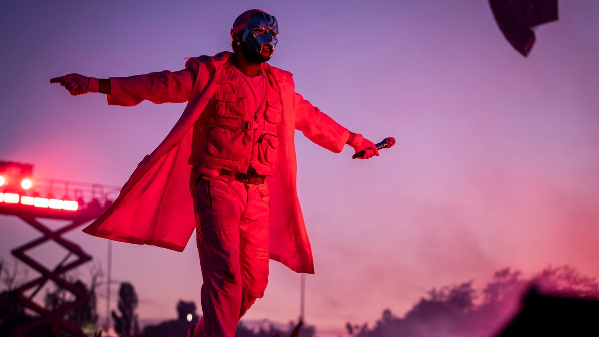 Abel Makkonen Tesfaye geht mit Gesichtsmaske, ausgebreiteten Armen in einem roten, offenen Mantel über Open-Air-Bühne. 