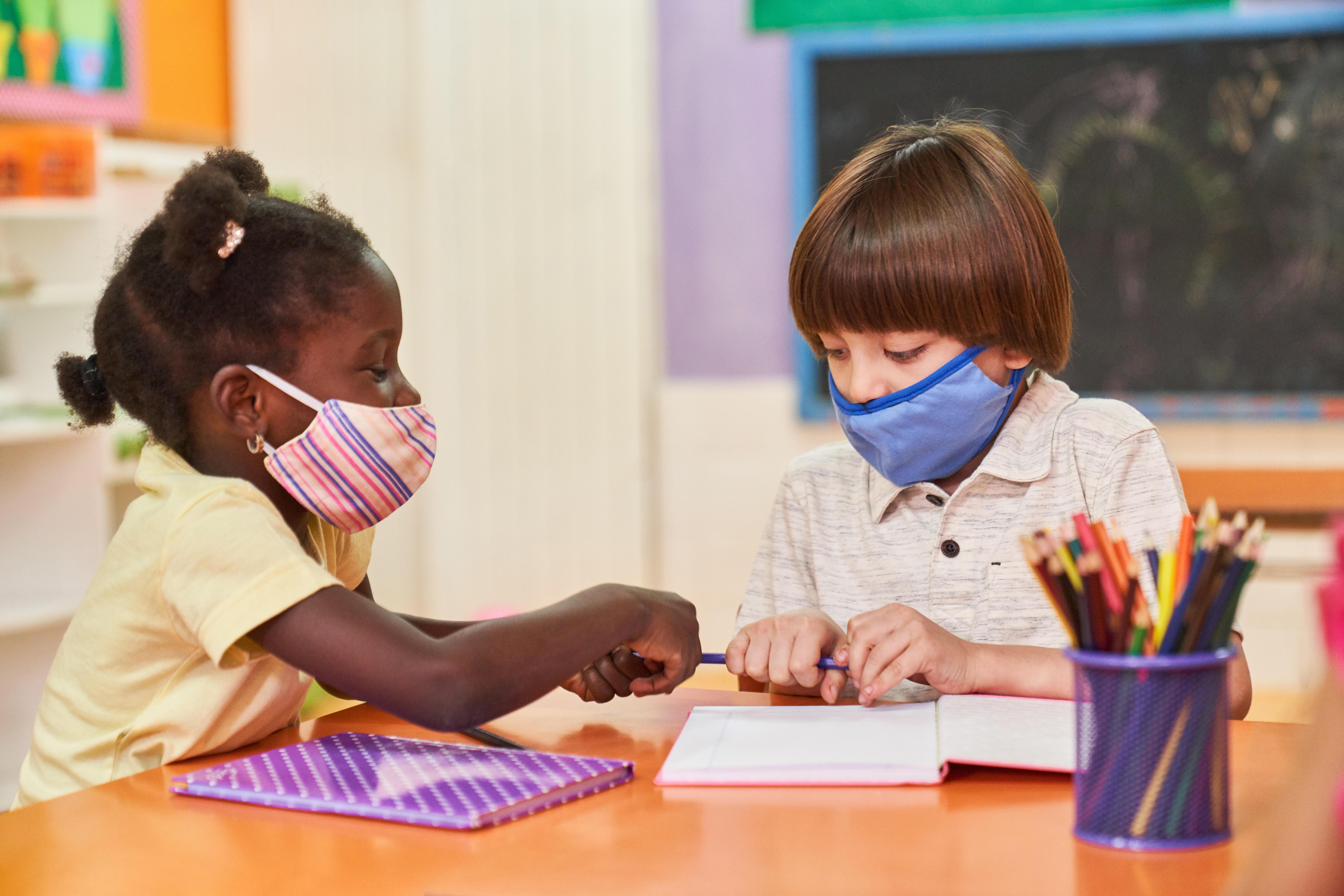 Zwei Kinder sitzen mit Mundschutz wegen Coronavirus-Pandemie an einem Tisch in der Grundschule oder Vorschule